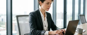 Сотрудница в деловом костюме работает за ноутбуком в офисе с панорамными окнами, на столе документы и кофе